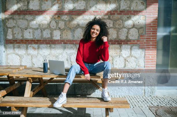 woman outdoors sitting on wooden table next to laptop, looking at camera. - red jumper stock pictures, royalty-free photos & images