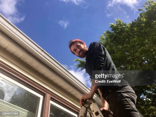 a man outside on a ladder cleaning the gutters of his house. - dakgoot stockfoto's en -beelden