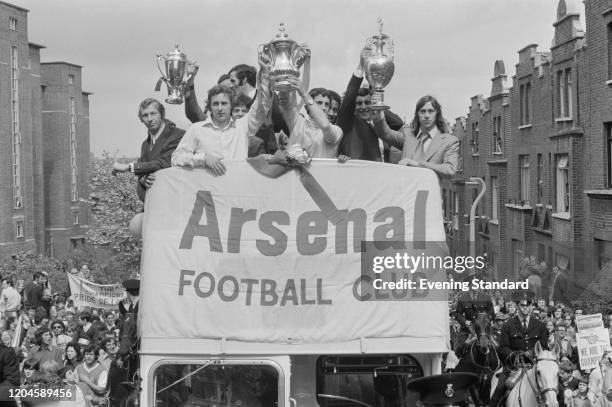 Members of the Arsenal FC team hold up trophies, including the League championship trophy and FA Cup trophy, as they parade in an open top bus...