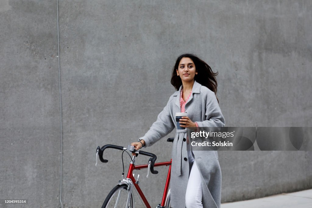 Young woman walking with bicycle, with reusable coffee cup in hand