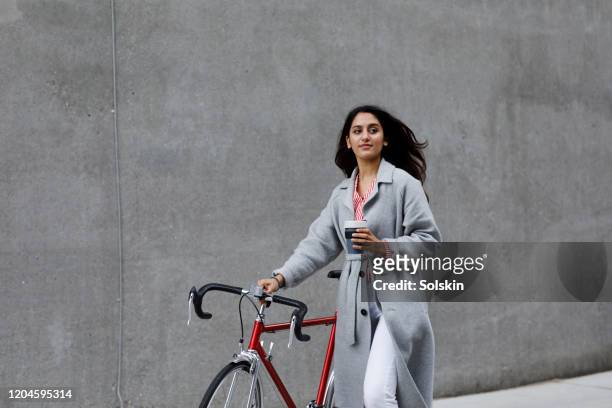 young woman walking with bicycle, with reusable coffee cup in hand - female likeness - fotografias e filmes do acervo