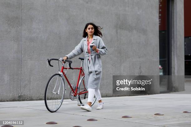 young woman walking with bicycle, with reusable coffee cup in hand - straat stockfoto's en -beelden