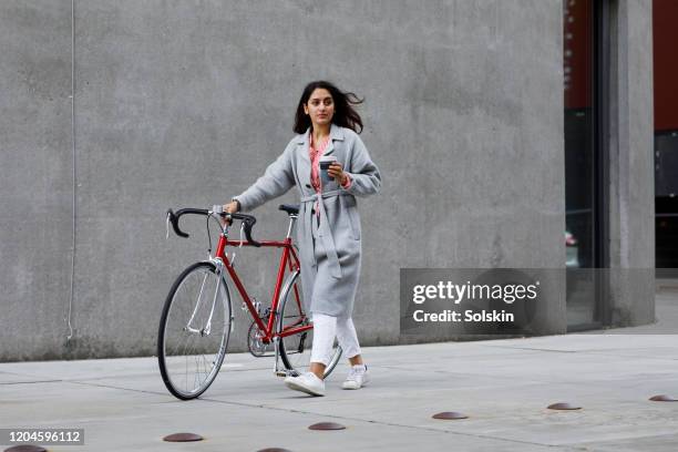 young woman walking with bicycle, with reusable coffee cup in hand - casaco cinzento imagens e fotografias de stock