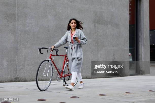 young woman walking with bicycle, with reusable coffee cup in hand - gray jacket imagens e fotografias de stock