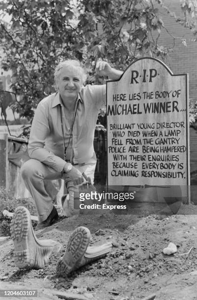 English film director and producer Michael Winner standing next to a fake tombstone which reads "RIP, Here Lies The Body Of Michael Winner, Brilliant...