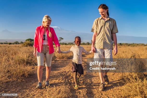 jovem casal caucasiano andando com seu filho africano adotado, monte kilimanjaro em segundo plano - missionário - fotografias e filmes do acervo