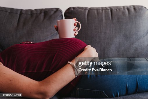 Portrait of young pregnant woman at home lying on the sofa and holding a cup on belly Portrait of young pregnant woman at home lying on the sofa and holding a cup on belly