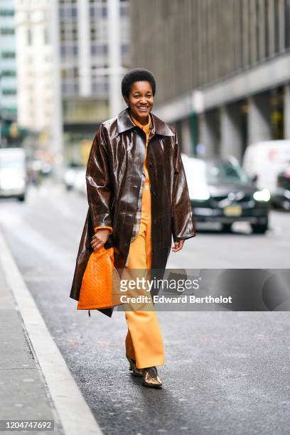 Tamu Mcpherson wears a brown leather shiny long coat, an orange knitted bag, orange flared pants, pointy shoes, outside Cinq a Sept, during New York...