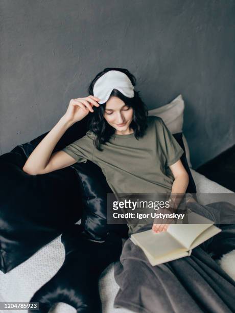 young woman lying on bed reading book - silk pillow stock-fotos und bilder