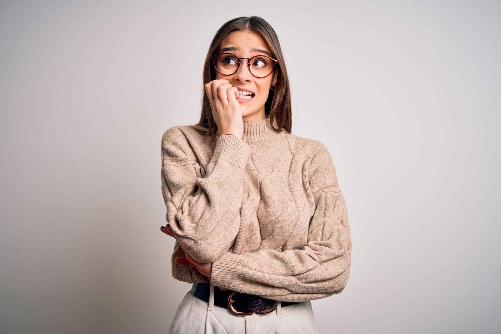 Young beautiful brunette businesswoman wearing casual sweater and glasses standing looking stressed and nervous with hands on mouth biting nails. Anxiety problem. Young beautiful brunette businesswoman wearing casual sweater and glasses standing looking stressed and nervous with hands on mouth biting nails. Anxiety problem.