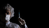 Female doctor in protection clothing holds syringe with vaccine in her hand on black background. Half face in shadow, dramatic emotion. Concept of virus protection, prevention of epidemics, treatment of diseases