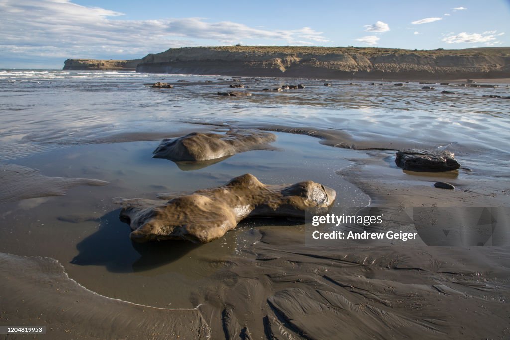 Wide shot of interesting rock formation on beach with reflections and sand dunes in background.