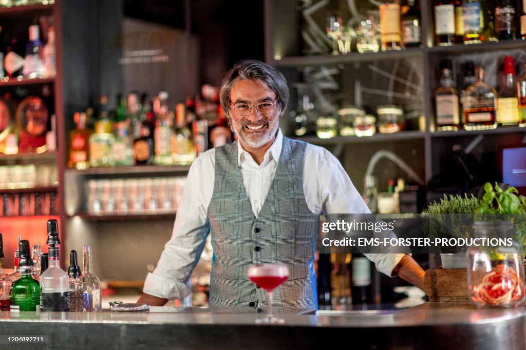 Smiling professional bearded barkeeper behind a counter in a hotel cocktail bar serving welcome drink