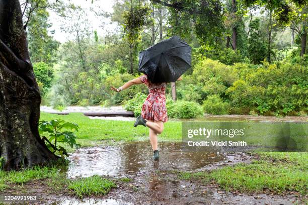 young female in a red dress and boots with a black umbrella dancing in a puddle under a tree - queensland storm stock pictures, royalty-free photos & images