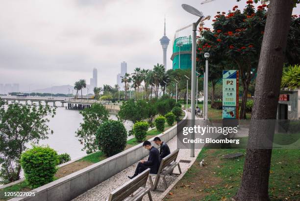 People wearing protective masks sit on a bench as Macau Tower stands in the distance in Macau, China, on Tuesday, March 3, 2020. Casinos in Macau,...