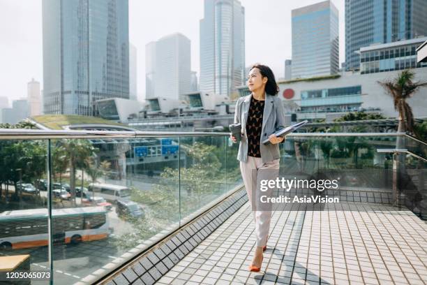 verticale de confiance femme d’affaires asiatique regardant loin et retenant des documents et ayant le café pour aller contre la scène de ville devant des immeubles modernes de bureau - hong kong business photos et images de collection