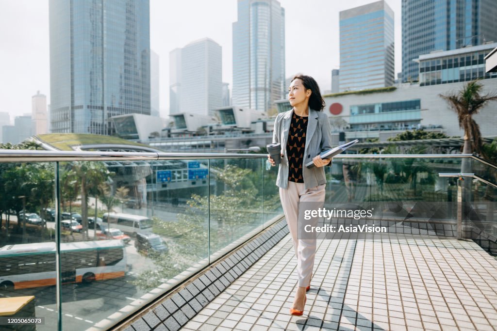 Portrait of confidence Asian businesswoman looking away and holding documents and having coffee to go against city scene in front of modern office buildings