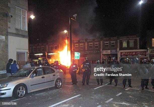 Riot police stand next to a burning shop which was set on fire by protesters over a fatal police shooting of a local man in Tottenham on August 6,...