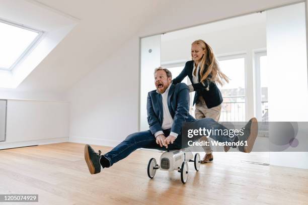 playful businesswoman pushing businessman on toy car in office - man riding toy car stock pictures, royalty-free photos & images