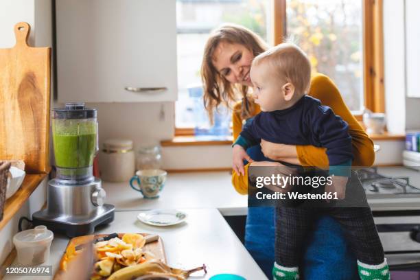 woman in the kitchen preparing healthy smoothie and holding her baby son - baby food stock pictures, royalty-free photos & images