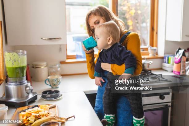 woman in the kitchen preparing healthy smoothie and holding her baby son - baby cup fotografías e imágenes de stock