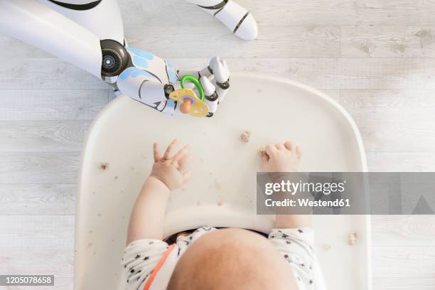 robot hand giving pacifier to baby boy sitting in high chair playing with bread crumps, top view - roboter hand stock-fotos und bilder