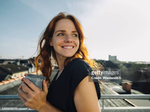 smiling redheaded woman having a coffee break on rooftop terrace - freude stock-fotos und bilder