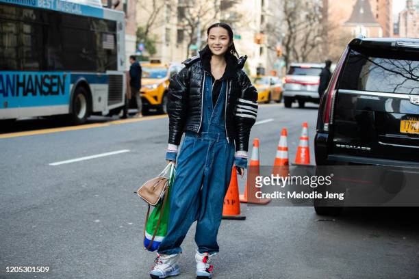 Model Xiao Wen Ju wears an asymmetrical cropped black puffer jacket, blue denim jumpsuit, Prrada bag, and baggy white sneakers on February 09, 2020...