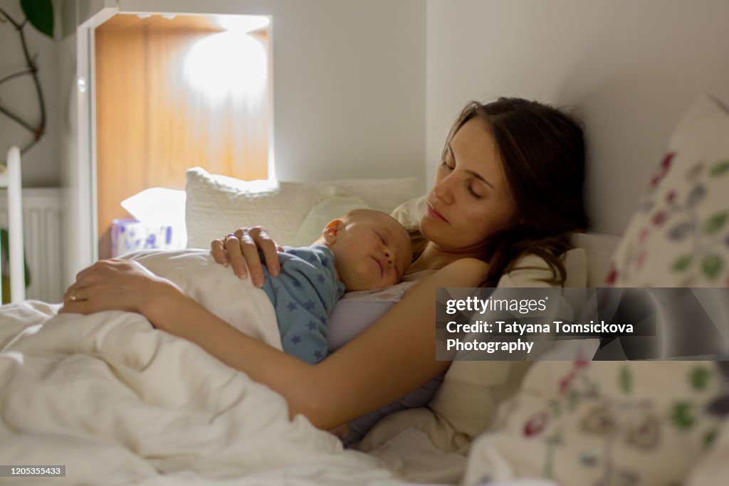 Young beautiful mother sleeping her newborn baby boy at night dim light. Mom breastfeeding infant