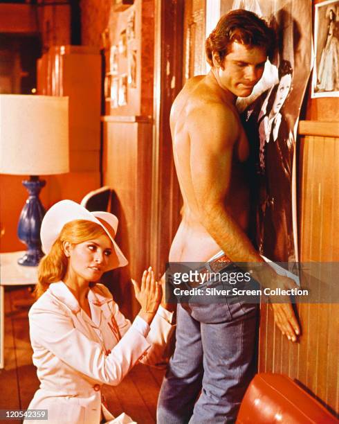 Raquel Welch, US actress, wearing a white wide-brimmed hat as she sits behind Roger Herren, US actor, who has his jeans down, exposing his buttocks...