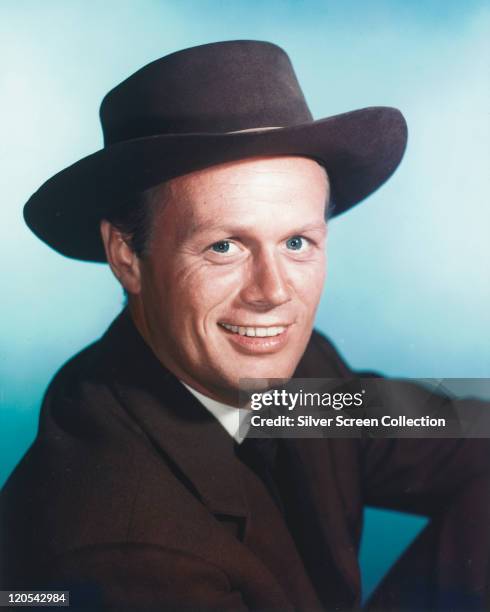 Richard Widmark , US actor, wearing a black wide-brimmed hat and black jacket smiling in a studio portrait, against a blue background, circa 1965.