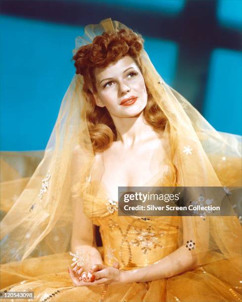 Rita Hayworth , US actress and dancer, wearing a yellow dress, with a yellow veil, in a studio portrait, against a blue background, circa 1945.