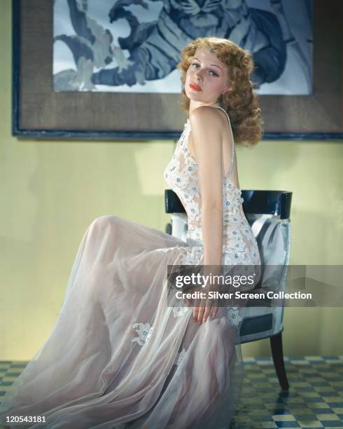 Rita Hayworth , US actress and dancer, wearing a long dress, with a white lace bodice, with floral motifs, and a white chiffon skirt, sitting in a...