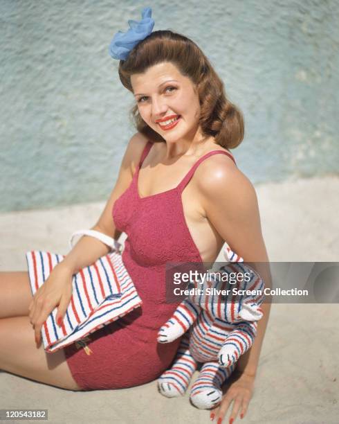Rita Hayworth , US actress and dancer, wearing a pink swimsuit and holding a red, white and blue striped towel in her lap and a red, white and blue...