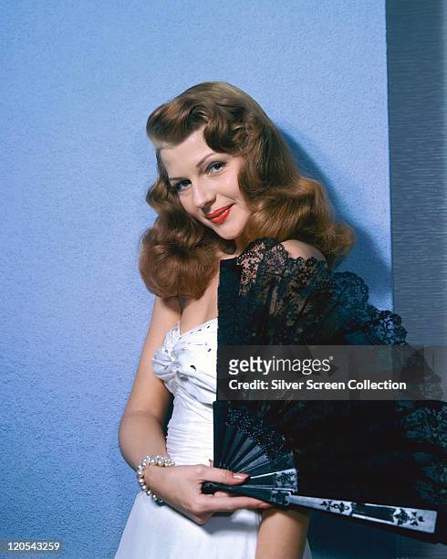 Rita Hayworth , US actress and dancer, wearing a white dress while posing with a black lace fan in a studio portrait, circa 1945.