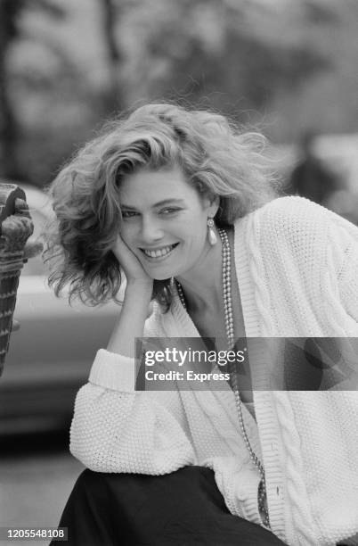 American actress Kelly McGillis, wearing a pearl necklace with white cardigan and dark skirt, smiles with her chin resting on her hand, in London,...