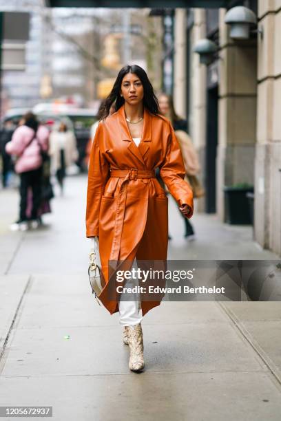 Guest wears an orange/brown leather long coat/dress, a Dior bag, white pants, snake print boots, during New York Fashion Week Fall Winter 2020, on...