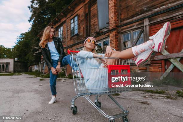 a long haired woman wearing a powder blue dress, pink woollen hat is chilling with her female friend, who is sitting on a shopping trolley, wearing a black leather jacket and blue jeans, in a derelict trading estate - pink shoe 個照片及圖片檔