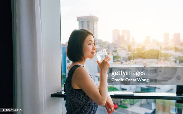 young woman drinking water while looking at view from the window - wasser trinken zu hause stock-fotos und bilder