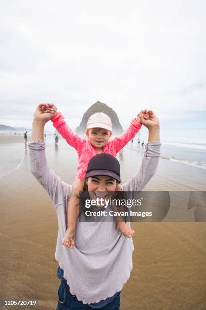 happy mom gives daughter shoulder ride at cannon beach. - cannon beach foto e immagini stock