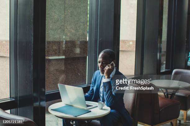 african businessman talking on his cell phone and looking at laptop - businessman in black suit stock pictures, royalty-free photos & images