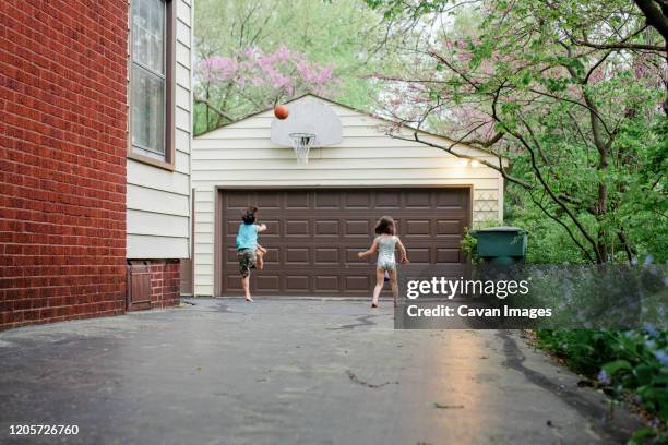 a boy leaps to shoot a basketball in driveway while sister watches on - garage driveway stock pictures, royalty-free photos & images