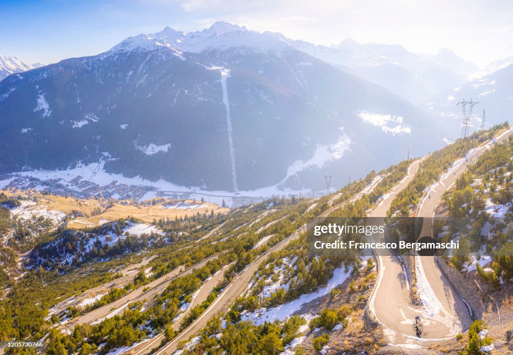 Road cyclist climbing hairpin bends up mountain pass in winter with snow.