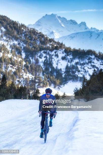 road cyclist rides a bycicle on a snowy and icy road in winter with a snowy mountain in the background. - road cycling stock pictures, royalty-free photos & images