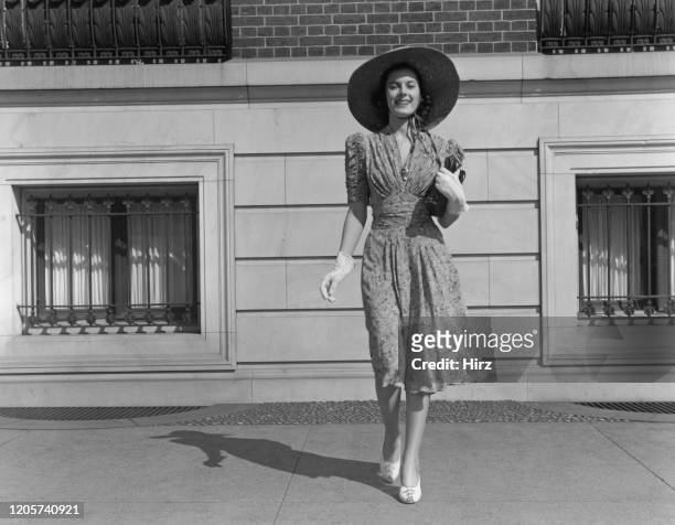 Woman wearing short-sleeved printed midi dress, daytime see-through gloves, sun hat with chin straps, and heeled shoes, circa 1950.