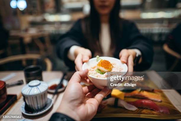 young woman passing a bowl of seafood donburi across table to a friend during lunch in a japanese restaurant - woman sushi stock-fotos und bilder