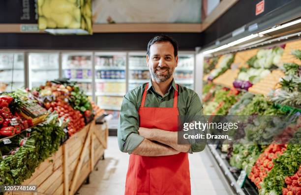 confident mature employee in supermarket, wearing red apron - salesman imagens e fotografias de stock