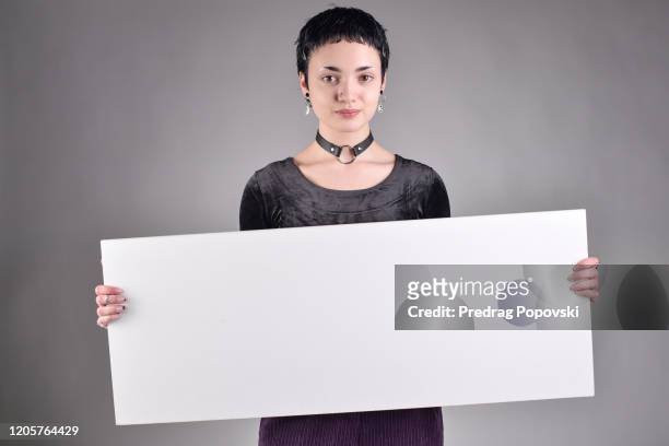cute short haired young modern woman with short hair in fashionable clothes holding blank white sign on studio background - frau mit plakat stock-fotos und bilder