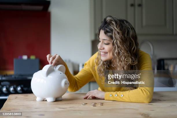cheerful young woman with curly hair at home saving coins into her piggybank - save money stock pictures, royalty-free photos & images