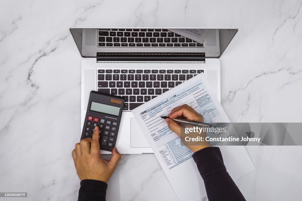 Overhead View of Hand Filling Up Tax Form and Using Calculator and Laptop